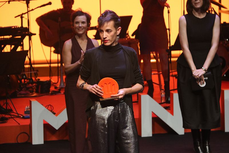 Blanca Llum Vidal, con chaqueta y pantalones plateados, sostiene un premio naranja en una ceremonia con músicos y público.