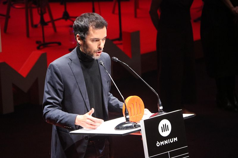 Discurso de aceptación del premio literario por la obra 'Elefants'. El ganador, con americana oscura y jersey negro, gesticula mientras habla desde un atril con el logo de la organización patrocinadora.