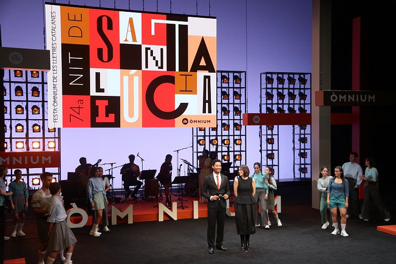 Escena de la 74ª Nit de Santa Llúcia: cartel con "SANTA LLÚCIA" y "OMNIUM", músicos, presentadores y actores en el escenario del Teatre Nacional de Catalunya.