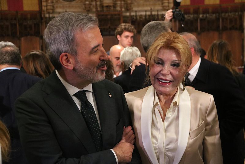 La Baronesa Thyssen-Bornemisza y el alcalde de Barcelona conversan y sonríen en un evento formal. Ella viste una chaqueta clara y él un traje oscuro.