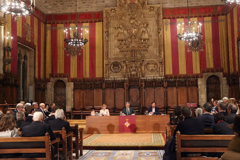 Presentación del futuro Museo Carmen Thyssen: rueda de prensa en salón antiguo con tres ponentes sentados en una mesa larga, frente a una audiencia en sillas de madera. Banderas con franjas rojas y amarillas decoran el espacio.