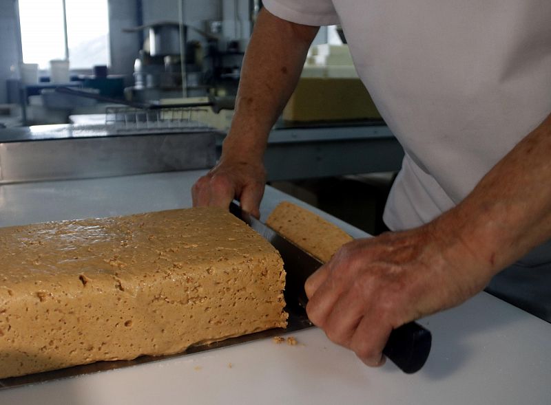 Corte de un bloque de turrón con almendras, de color marrón claro y textura densa, usando un cuchillo de mango negro sobre una superficie blanca.