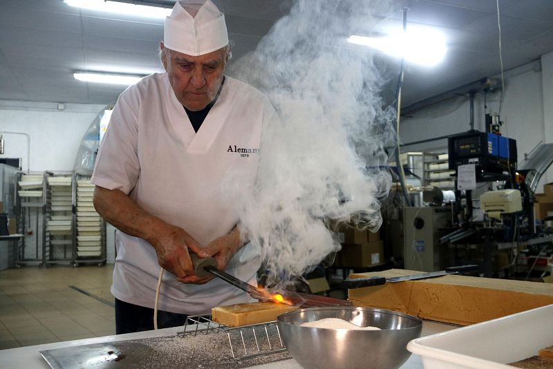 Trabajador con uniforme quema turrón con soplete, creando humo y caramelizando el azúcar.  Se observa azúcar y cuchillo cerca.