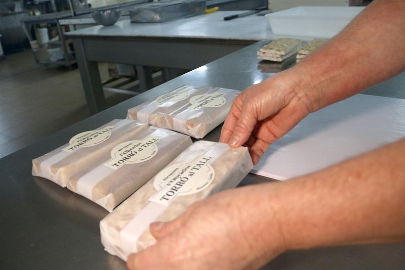Empaquetado de barras de turrón 'L’Obraor TORRÓ al TALL', envueltas en papel pergamino y con etiqueta circular.  Una persona manipula una barra sobre una superficie blanca en un taller.