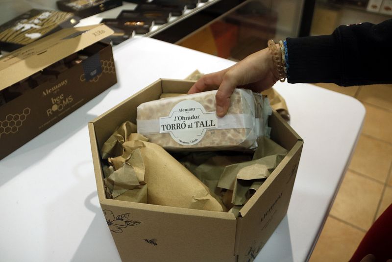 Mano colocando turrón Alemany en caja hexagonal de cartón. El envoltorio muestra 'TORRÓ al TALL' y el logo de la marca.  Caja rectangular de Alemany al fondo.