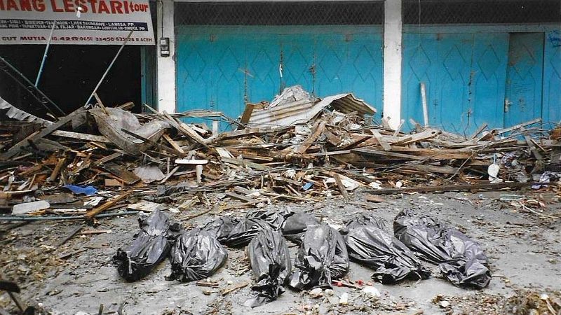 Bolsas para cadáveres y escombros cubren el suelo tras el devastador tsunami. Una pared parcialmente destruida se alza al fondo, mostrando la magnitud de la destrucción.