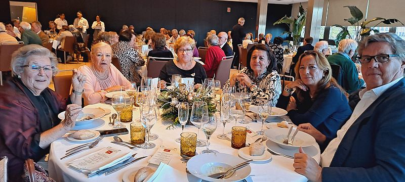 Almuerzo navideño festivo: siete personas alrededor de una mesa con mantel blanco y centro de mesa navideño en un salón decorado.