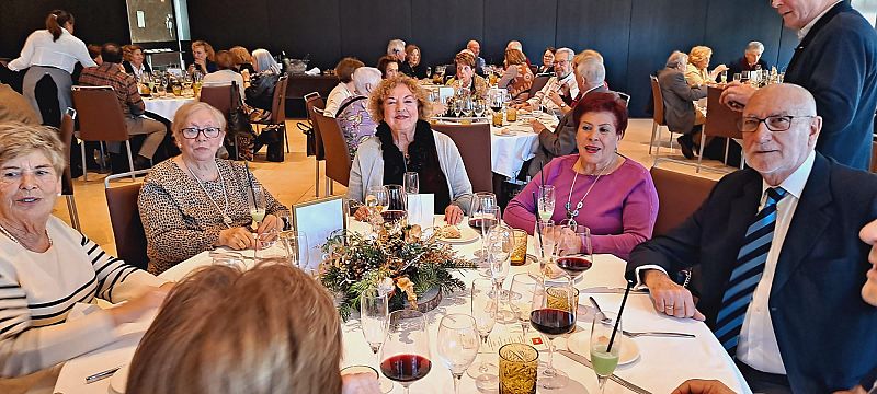 Comensales en un almuerzo navideño formal, con mesas decoradas y ambiente festivo.  Se aprecian platos, copas y decoración navideña.