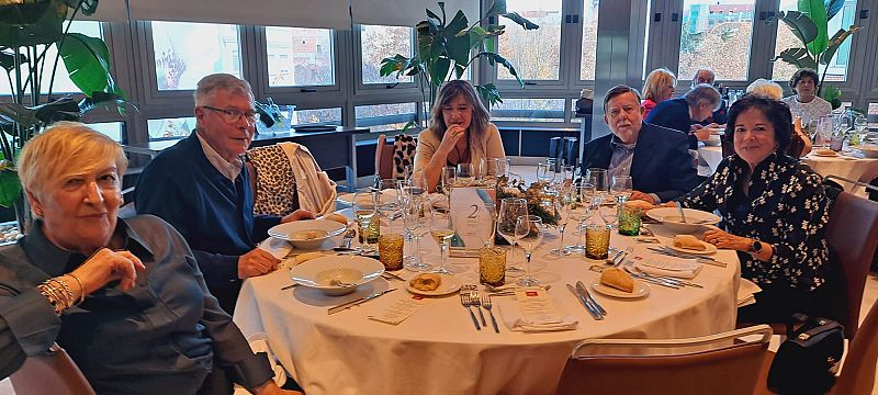 Almuerzo navideño de seis personas alrededor de una mesa formalmente ambientada, con decoración navideña y abundante luz natural.