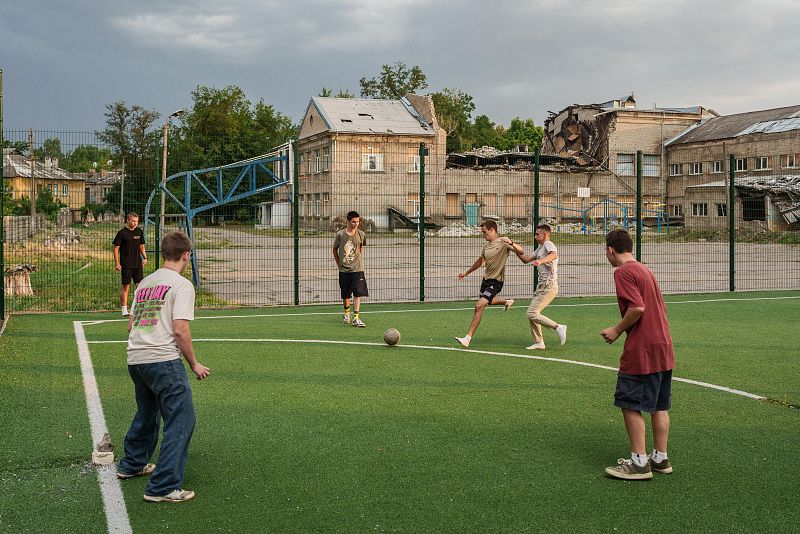 Jóvenes jugando al fútbol en una cancha con un edificio bombardeado al fondo.  Contrasta la alegría del juego con la destrucción del entorno.