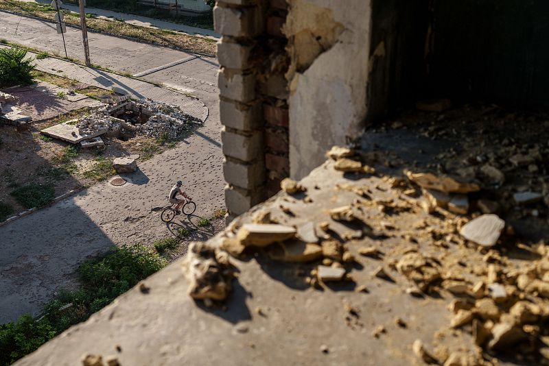 Escombros y un cráter en una calle dañada. Una persona en bicicleta pasa cerca de los restos de un ataque. Tonos grises y marrones predominan en la imagen.