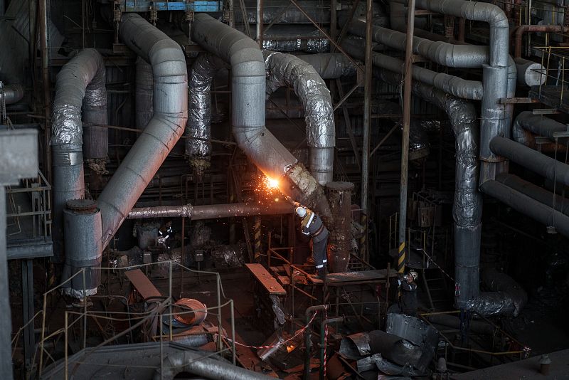 Interior deteriorado de una central eléctrica; trabajador con equipo de protección soldando o cortando una tubería; escombros en el suelo indican desmantelamiento.