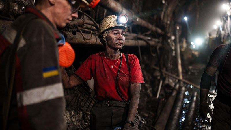 Minera ucraniana con casco y linterna, rostro manchado de polvo, expresión seria, trabajando en mina oscura con otros mineros.  Uniformes oscuros, bandera ucraniana visible.