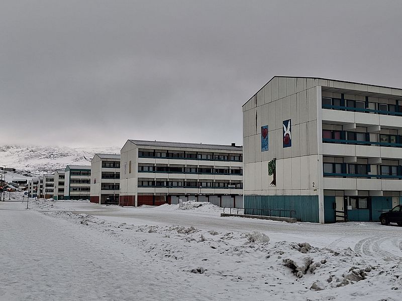 Edificios blancos de varios pisos con detalles verdes y murales en un paisaje nevado. Nieve acumulada y un coche negro visible.