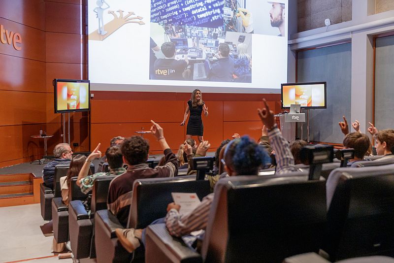 Presentadora en sala de conferencias de RTVE dirigiéndose a estudiantes de 4º ESO sobre tecnología y empresa.  Imágenes de programación en pantalla.  Participación activa del público.