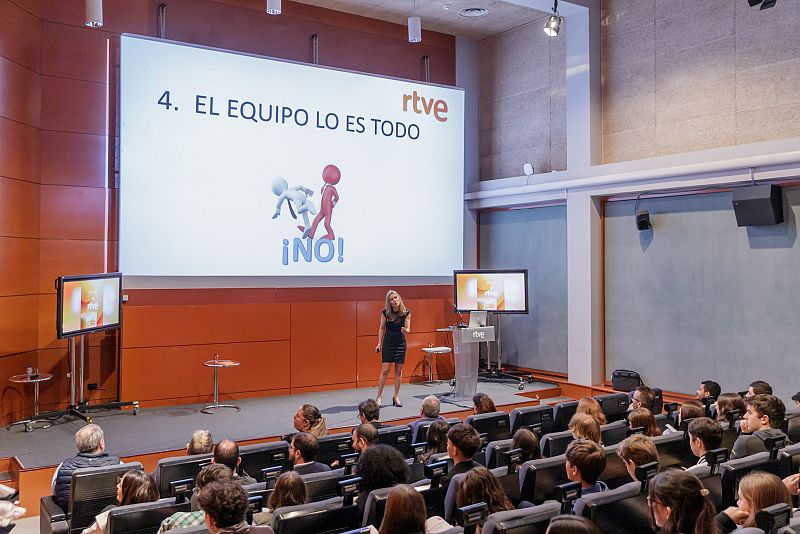 Presentación en sala de conferencias con pantalla mostrando el logo de RTVE y el texto "4. EL EQUIPO LO ES TODO" y "¡NO!".  Presentadora en traje negro ante público joven.