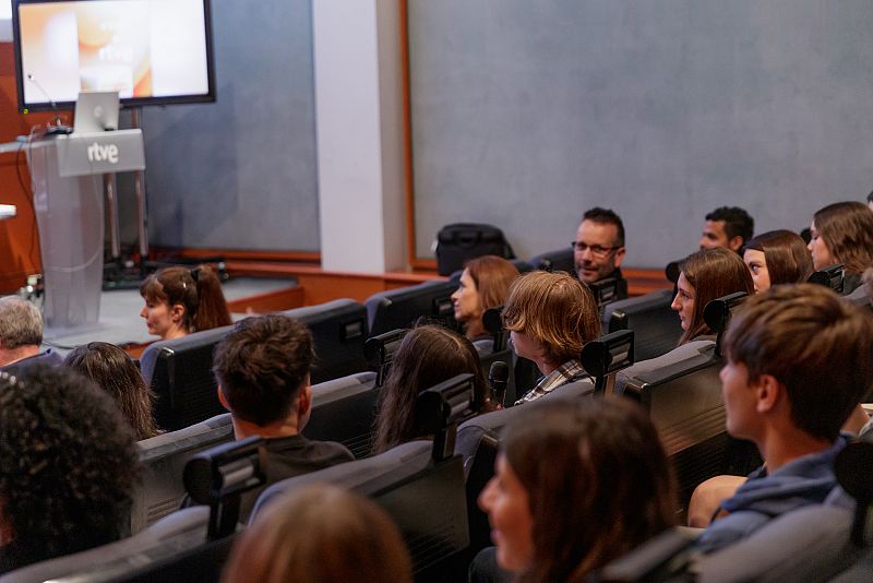 Auditorio con público asistente a una presentación.  Pantalla grande mostrando diapositivas y atril con logo de RTVE.  Asisten estudiantes y adultos.