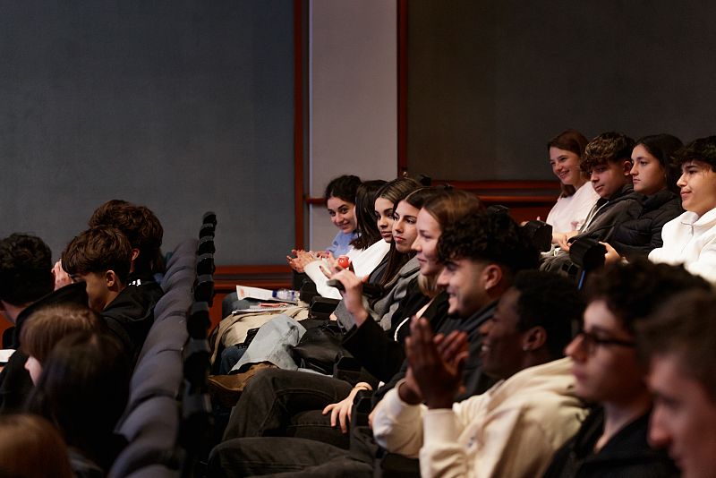 Jóvenes sentados en un auditorio, algunos aplauden, otros miran hacia el frente.  Ambiente de participación en un evento.