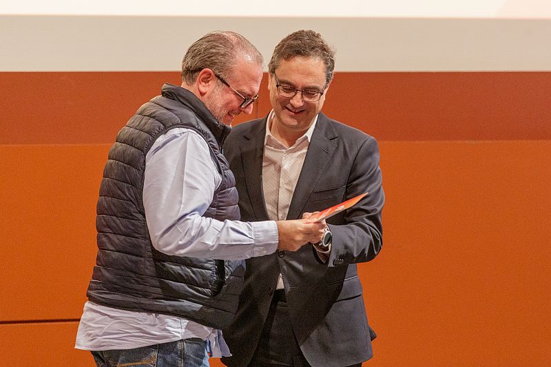 Intercambio de un objeto (posiblemente un diploma) entre dos hombres en un ambiente formal y positivo; uno viste traje y el otro camisa azul y chaleco.