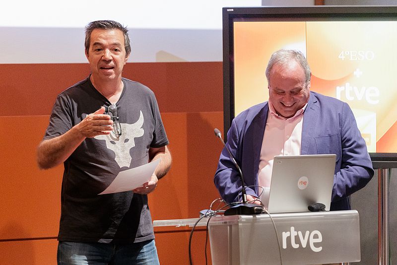 En una presentación, un hombre con camiseta negra habla mientras otro, con chaqueta azul, revisa un portátil RTVE sobre un atril con el mismo logo.  Se observa una pantalla con "4º ESO + rtve".