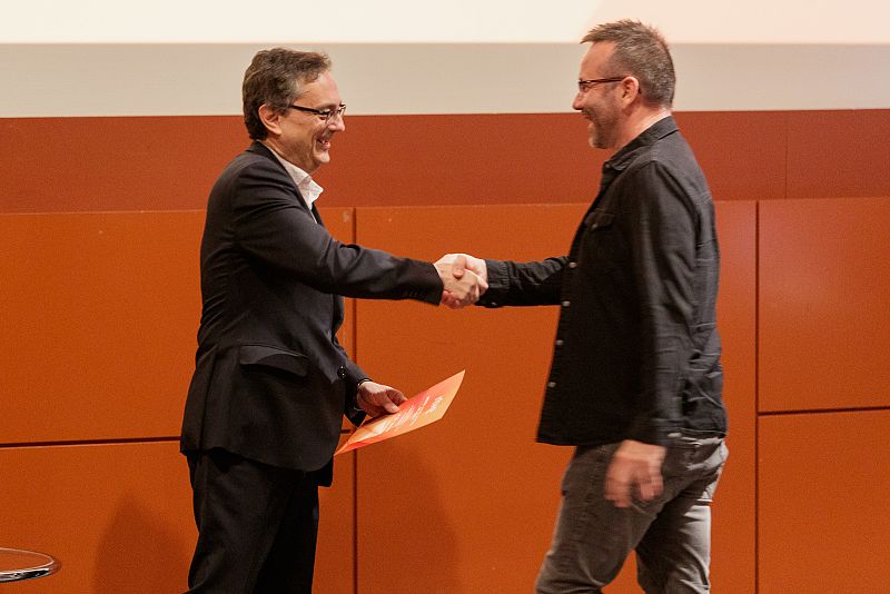 Dos hombres, uno en traje oscuro con gafas y un documento naranja, el otro con camisa oscura y pantalones grises, se dan la mano sonriendo en un evento con fondo naranja.