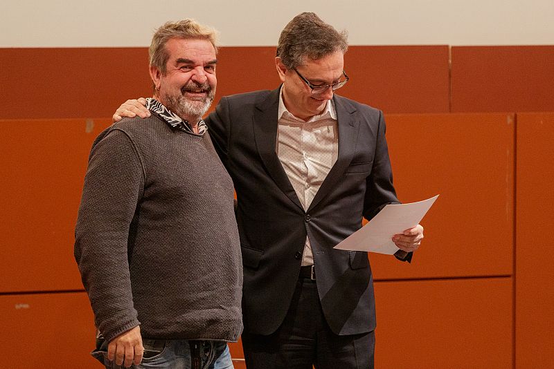 Dos hombres, uno en traje con papeles y otro con jersey gris, sonríen en una sala con paneles naranjas de fondo.