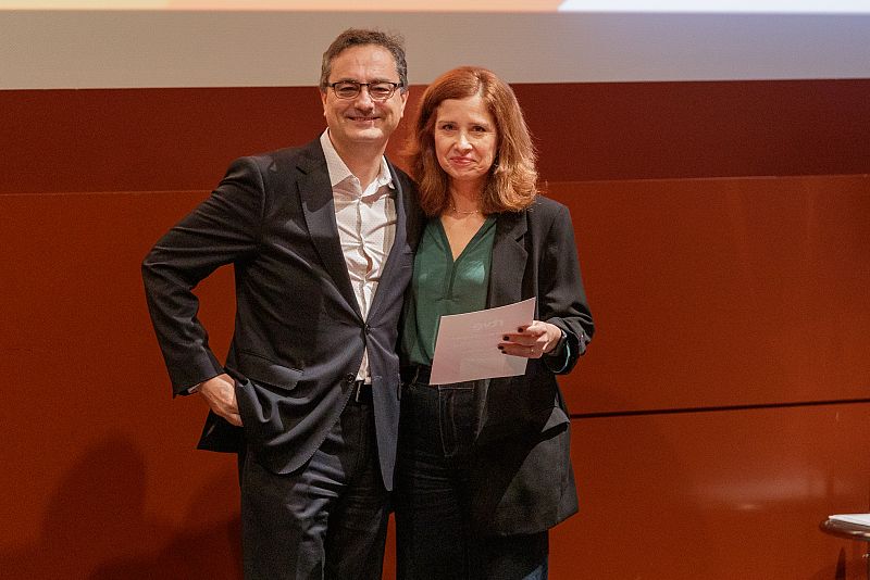 Dos personas, formalmente vestidas, en una gala. La mujer sostiene un papel. Fondo anaranjado.