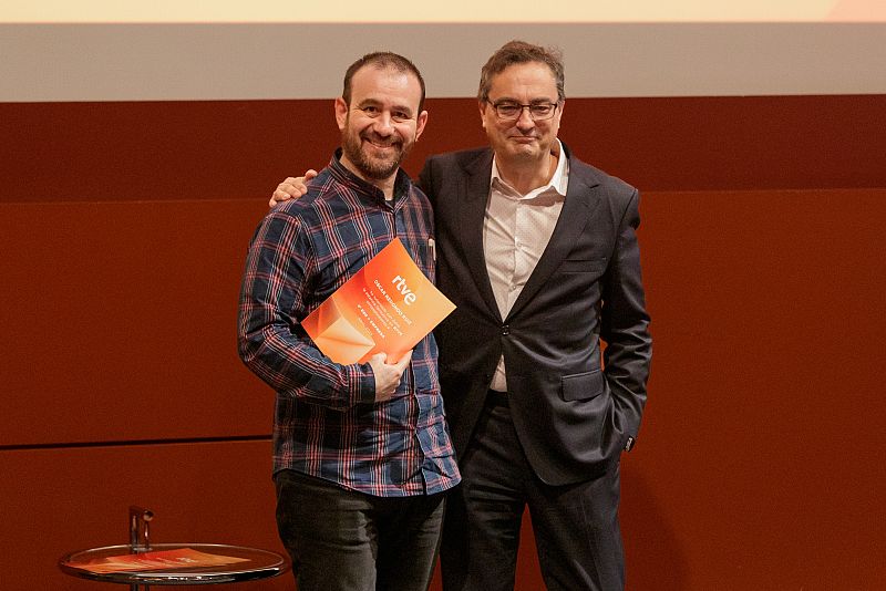 Dos hombres, uno con camisa a cuadros y un documento con logo RTVE, el otro con traje, posan en una gala con fondo rojizo.