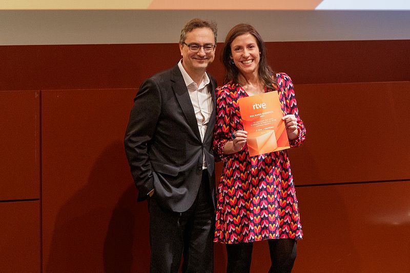 Entrega de un diploma con el logo de RTVE en una gala. Una mujer con vestido floral y un hombre con traje participan en la ceremonia.