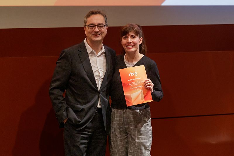 Gala de bienvenida: hombre en traje y mujer con chaqueta negra y diploma con logo RTVE, fondo granate.