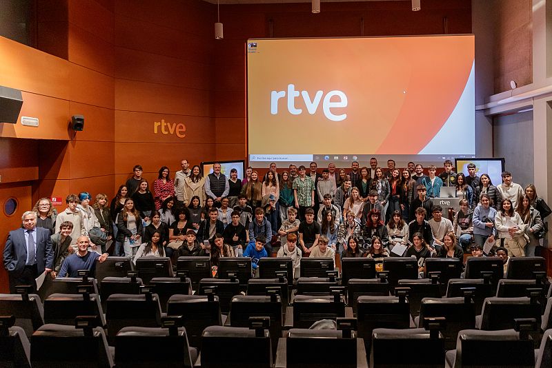 Grupo de estudiantes de 4º ESO y adultos en una sala de RTVE, durante una gala de bienvenida.  Se observa el logo de RTVE en una pantalla y en la pared.  Ambiente festivo.