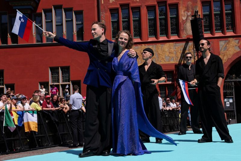 Delegación eslovena en la Alfombra Turquesa de Eurovisión 2025: un hombre con chaqueta azul y una mujer con vestido largo azul posan con otros dos acompañantes. Multitud de fondo.