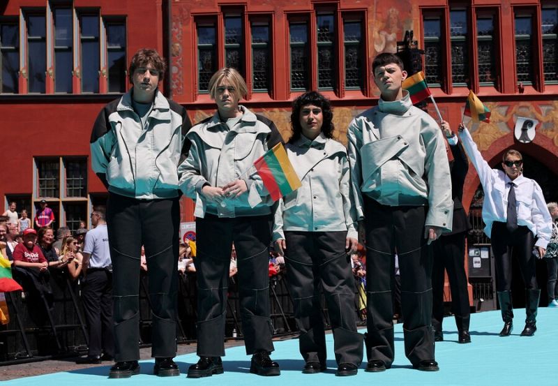 Grupo de cuatro personas con atuendos grises y negros posan en alfombra turquesa, sosteniendo banderas lituanas. Al fondo, otra persona con camisa blanca y corbata agita una bandera mayor.