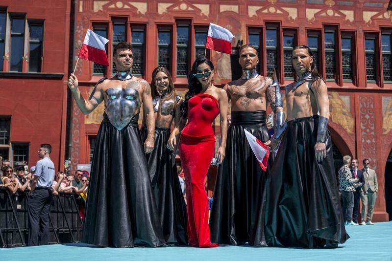 Cinco personas, una con vestido rojo y otras con trajes oscuros, posan en una alfombra turquesa. Dos portan banderas polacas, sugiriendo una representación de Polonia.