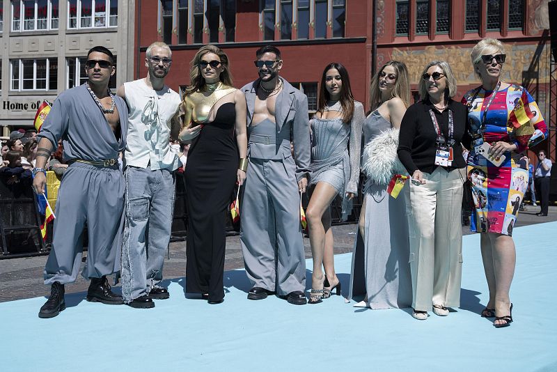 Grupo de ocho personas con atuendos elegantes posa en alfombra turquesa, frente a edificios. Algunas llevan banderas españolas.