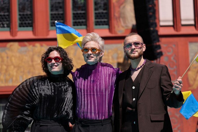 Grupo de tres personas con atuendos elegantes y gafas de sol, dos portando banderas ucranianas, posando en una alfombra turquesa.