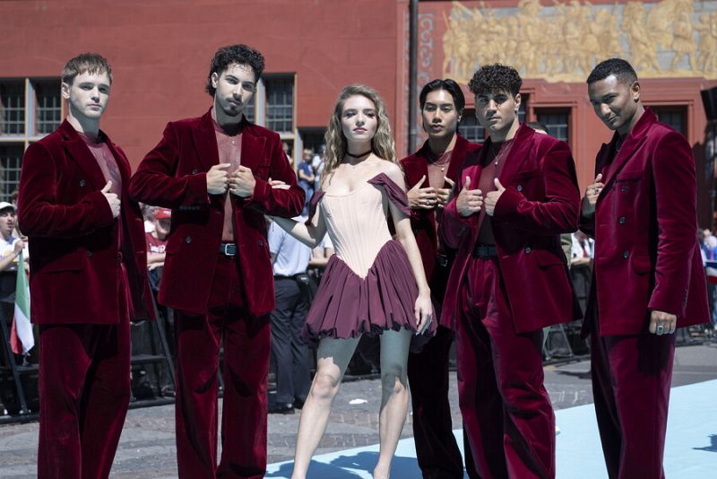 Grupo de cinco jóvenes, cuatro hombres en trajes granate y una mujer con vestido burdeos, posan juntos sobre una alfombra turquesa. Fondo con público y mural.