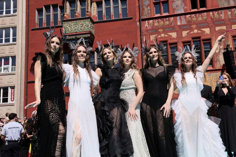 Cinco mujeres con vestidos largos y tocados plateados posan en una alfombra. Dos lucen vestidos blancos, dos negros y una beige. Fondo con edificio de ladrillo rojo y público.