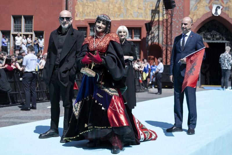 Cantante albanesa en la Alfombra Turquesa de Eurovisión 2025 con vestido rojo y negro, sosteniendo un premio dorado y acompañada por su delegación.