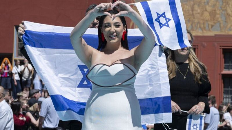 Representante de Israel en Eurovisión 2025 en la Alfombra Turquesa, con vestido blanco y gesto de corazón. Banderas de Israel al fondo.