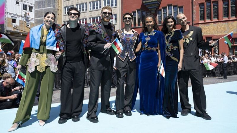 Grupo musical azerbaiyano posa en alfombra turquesa con atuendos tradicionales y banderas. Evento público, posiblemente Eurovisión.