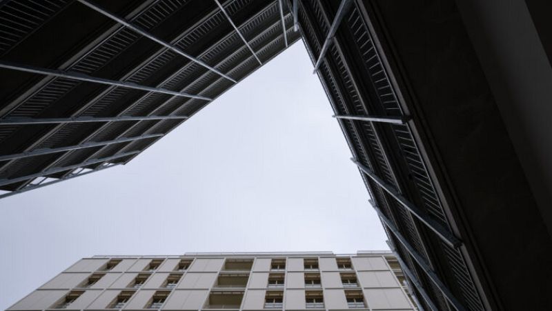 Patio interior con dos edificios de balcones metálicos oscuros enmarcando un tercer edificio más alto al fondo, bajo un cielo gris claro.