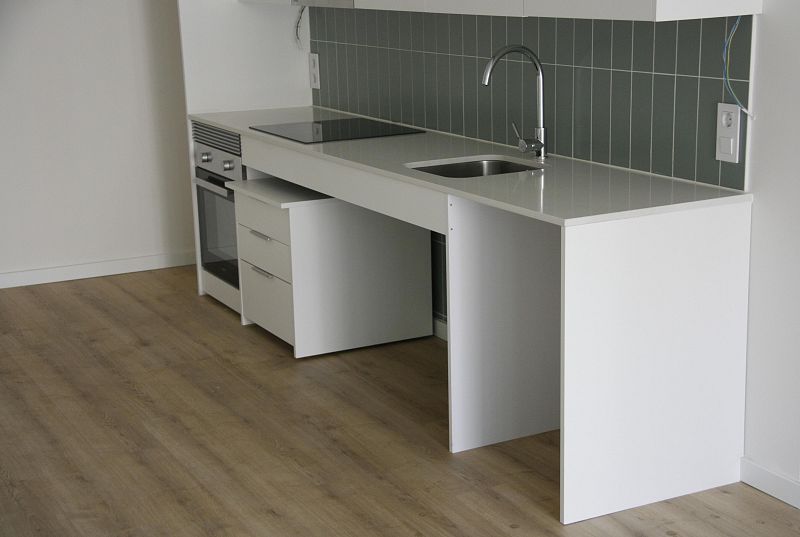 Cocina moderna minimalista: encimera blanca, placa vitrocerámica, horno integrado, azulejos grises verdosos y suelo de madera clara.