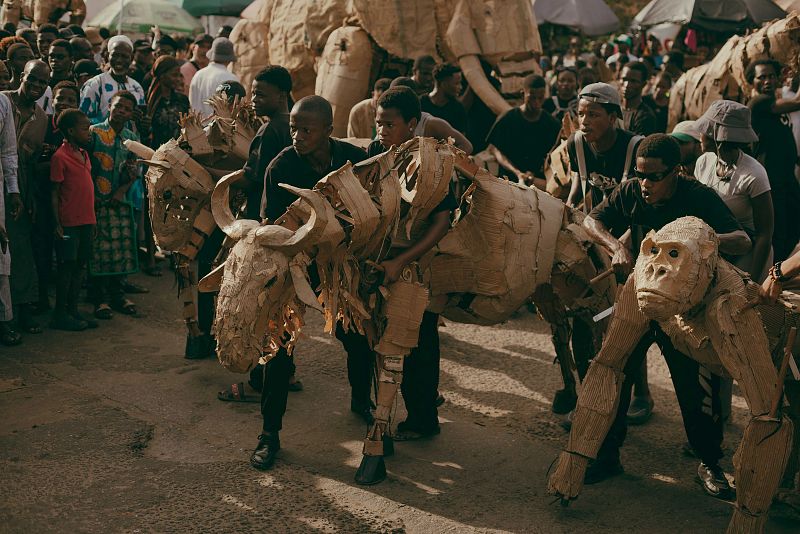 Actuación callejera con figuras de cartón de animales (antílope y gorila) manipuladas por varias personas ante una multitud en un entorno urbano soleado.