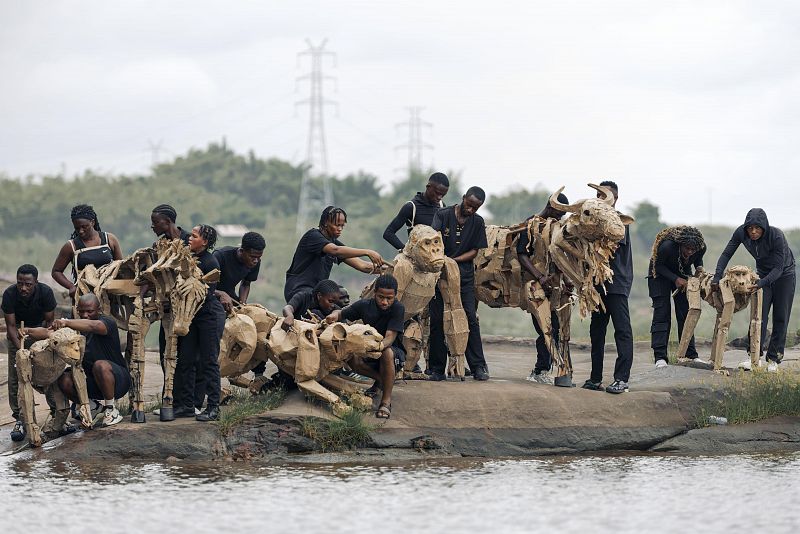 Marionetas de animales africanos, construidas con materiales reciclados, son manipuladas por titiriteros vestidos de negro a orillas de un río.  Posible espectáculo al aire libre.