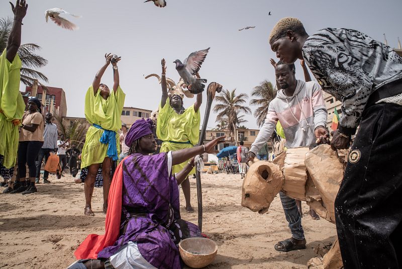 Actuación en una playa: persona con ropa morada y turbante interactúa con figura de cartón; otros con ropa verde levantan las manos; palomas volando; entorno costero urbano.