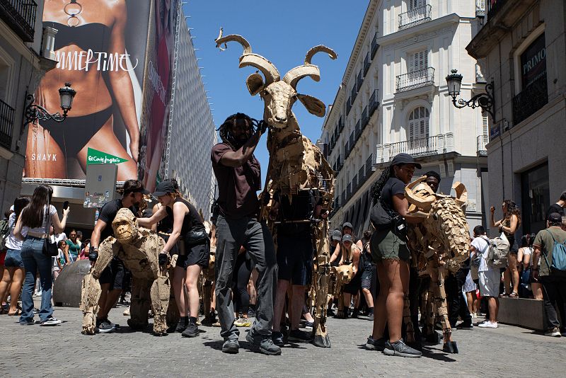 Marionetas gigantes, incluyendo una cabra y un gorila, son manipuladas en una calle peatonal. Un anuncio con una mujer en bikini y el texto "SUMMER TIME" es visible en el fondo.