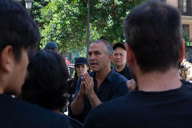 Director artístico dirigiendo a un grupo, posiblemente titiriteros, antes de un desfile al aire libre.  Se observa a personas escuchando atentamente sus instrucciones.