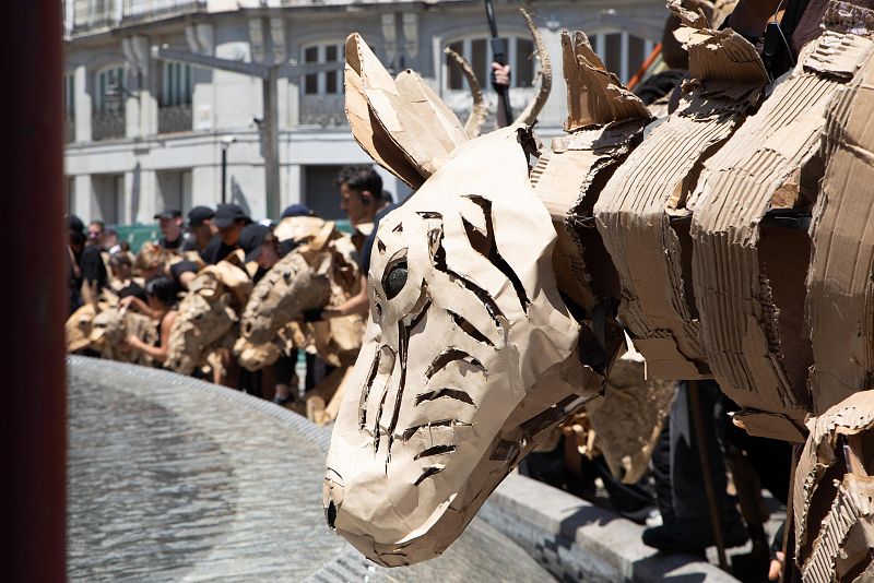 Marioneta de cartón con forma animal (posiblemente cebra) junto a una fuente.  Al fondo, más marionetas y personas en una actuación.