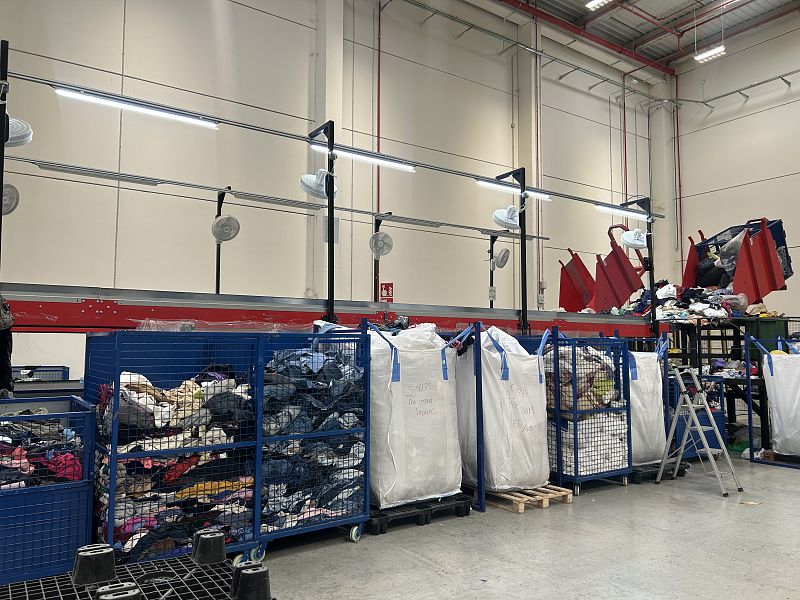 Interior de una planta de reciclaje: contenedores azules con ropa, cinta transportadora roja, carros metálicos y sacos blancos llenos de prendas. Iluminación y ventilación industrial.